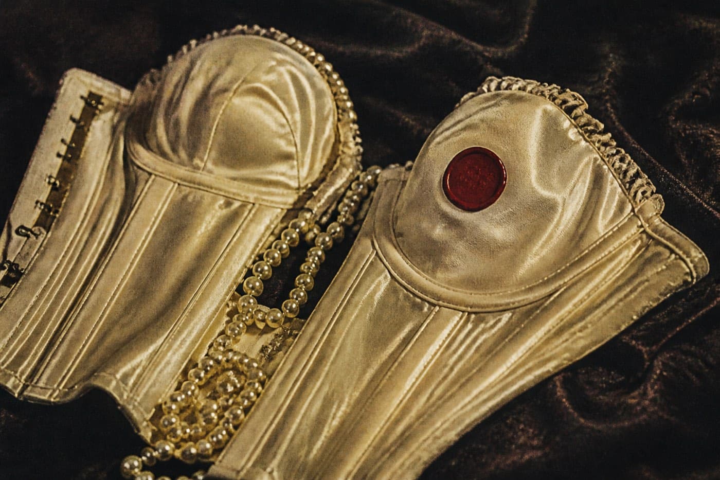 A cream satin corset on velvet with a spill of pearls across the cups, closed with a red wax seal.