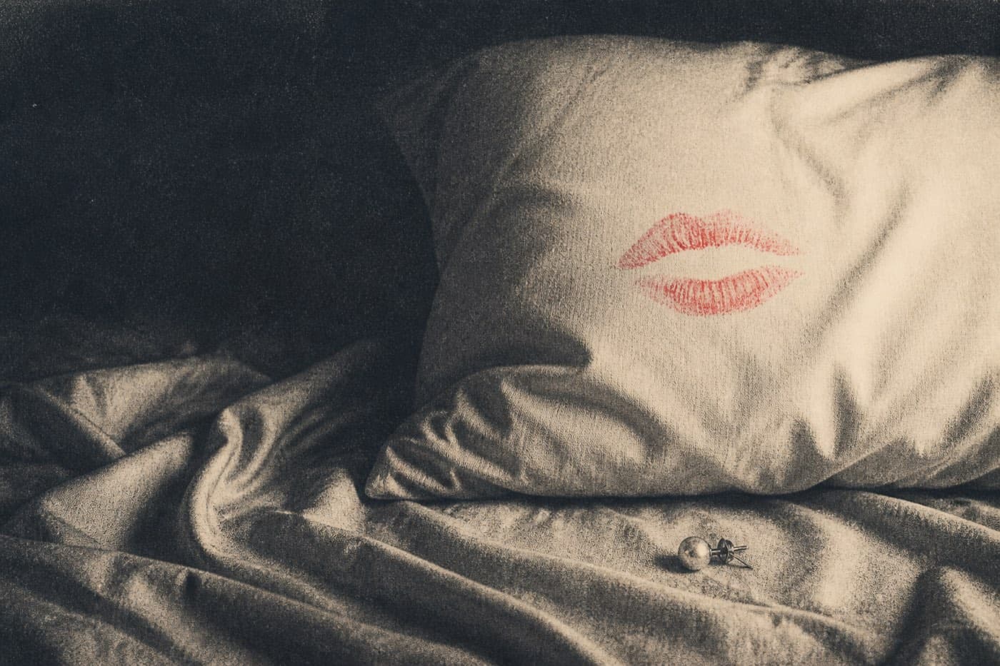 A cream pillow with a lipstick smear across it, one pearl earring left on rumpled sheets.